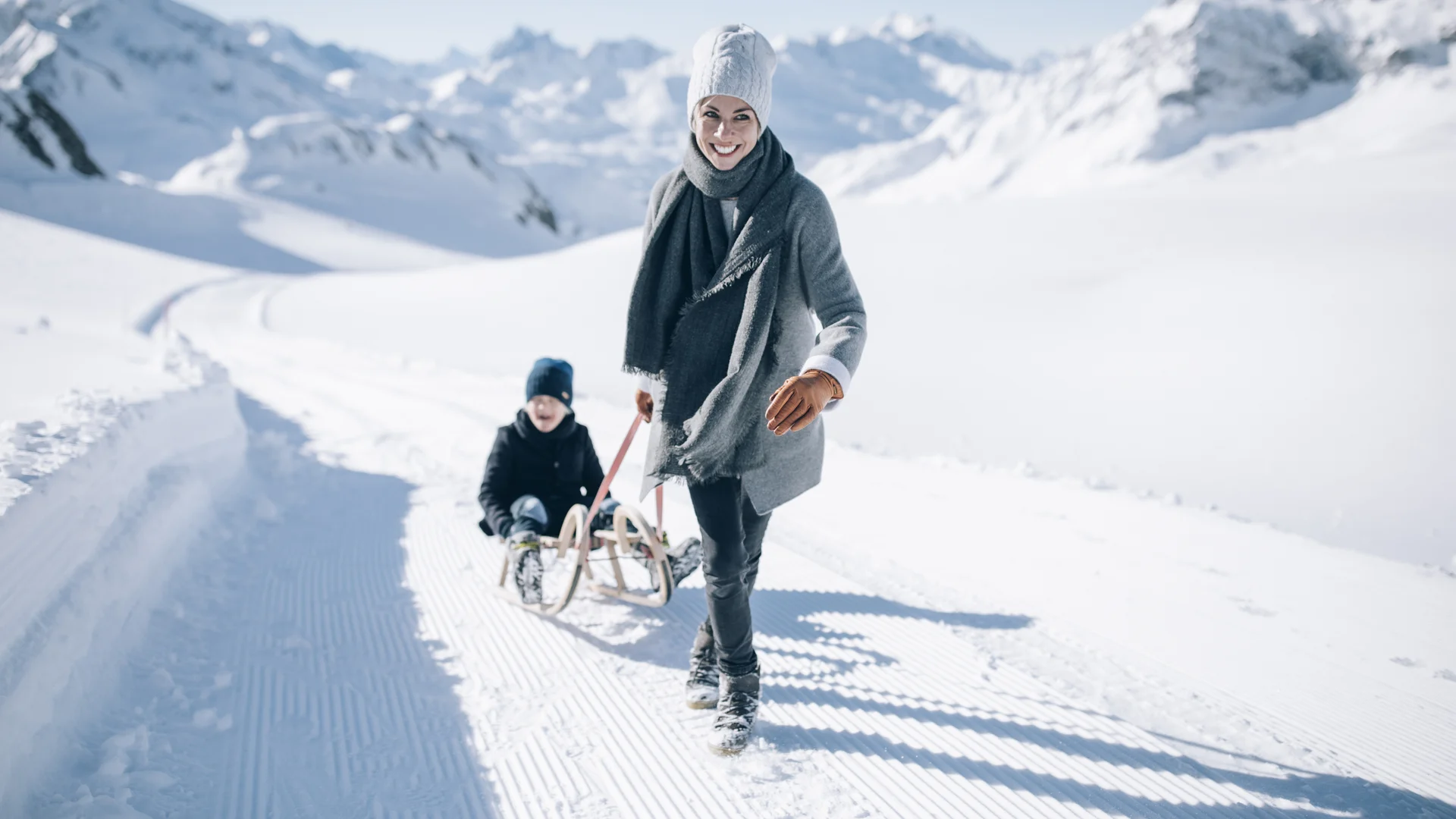 Hotel in Lech am Arlberg mit Ski-in/Ski-out | Hotel Panorama Frau und Kind mit Schlitten auf einem präparierten Winterweg vor verschneiten Bergen